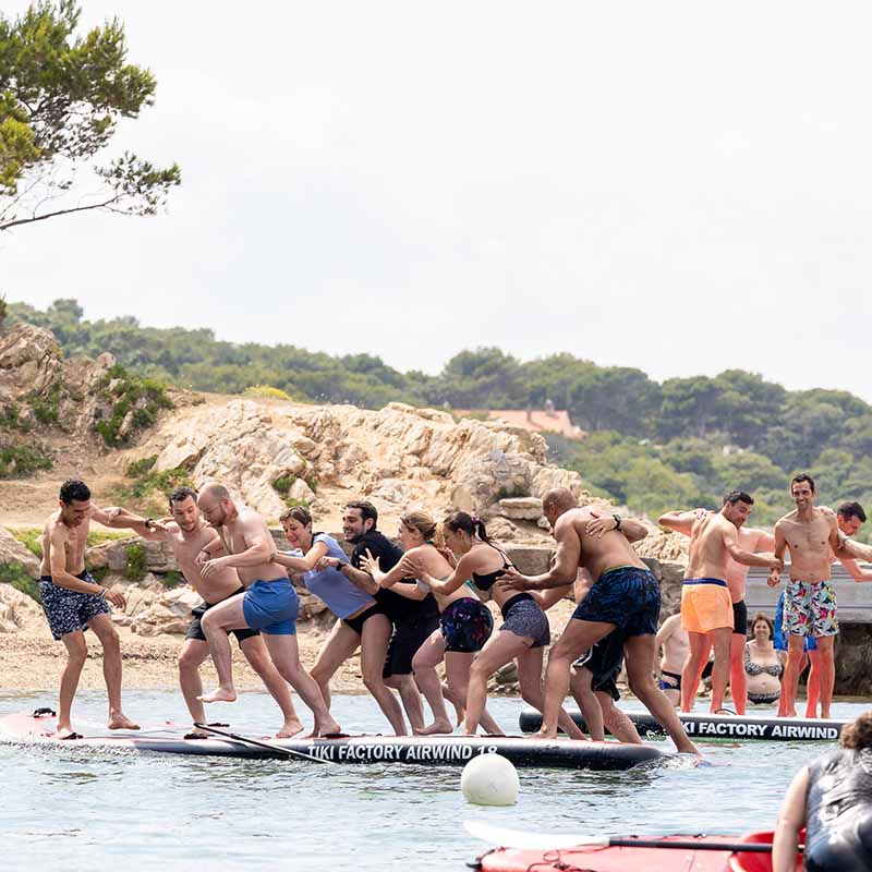 Photo team building Marseille : collaborateurs en maillot de bain s'amusant sur une planche de paddle géante en pleine mer.