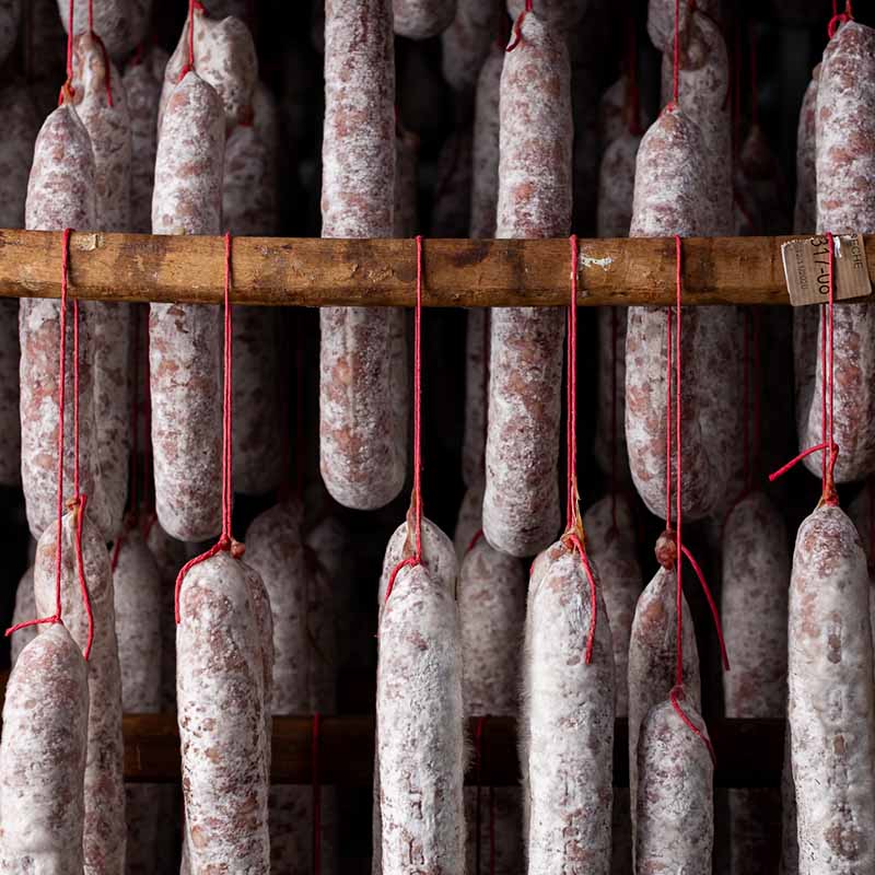Photo artisan Marseille : alignement de saucissons artisanaux suspendus par des fils rouges dans un séchoir traditionnel.