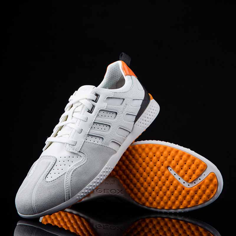 Photographie packshot de baskets Geox blanches et grises avec semelle orange dynamique sur fond noir, reflet élégant.