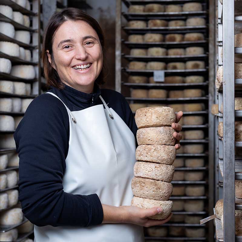 Portrait d'une productrice de fromage souriante tenant une pile de tommes artisanales dans son séchoir près de Marseille.
