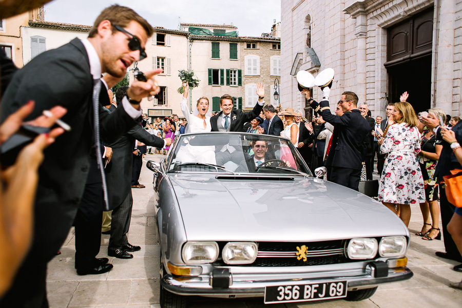 Un couple de mariés tout juste unis quittant l'église en voiture ancienne cabriolet grise sous les acclamations des invités à Marseille.