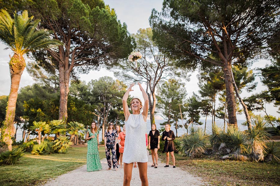 Mariée en robe courte scintillante jetant son bouquet de fleurs à ses amies lors d'un mariage en extérieur à Marseille.