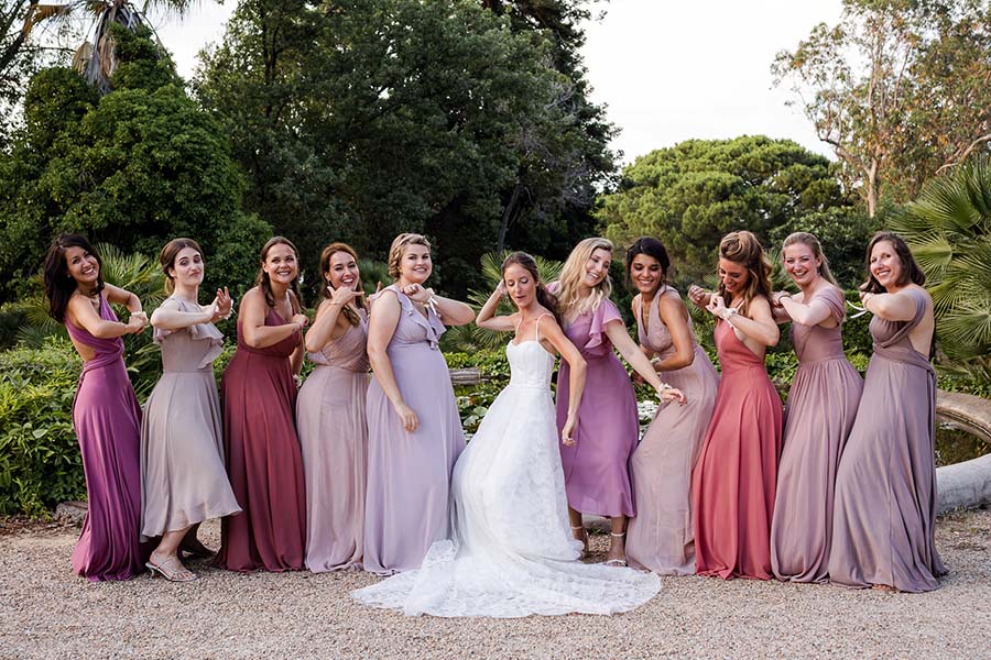 La mariée entourée de ses dix demoiselles d'honneur portant des robes dégradées de rose et mauve, posant de façon fun dans un jardin à Marseille.