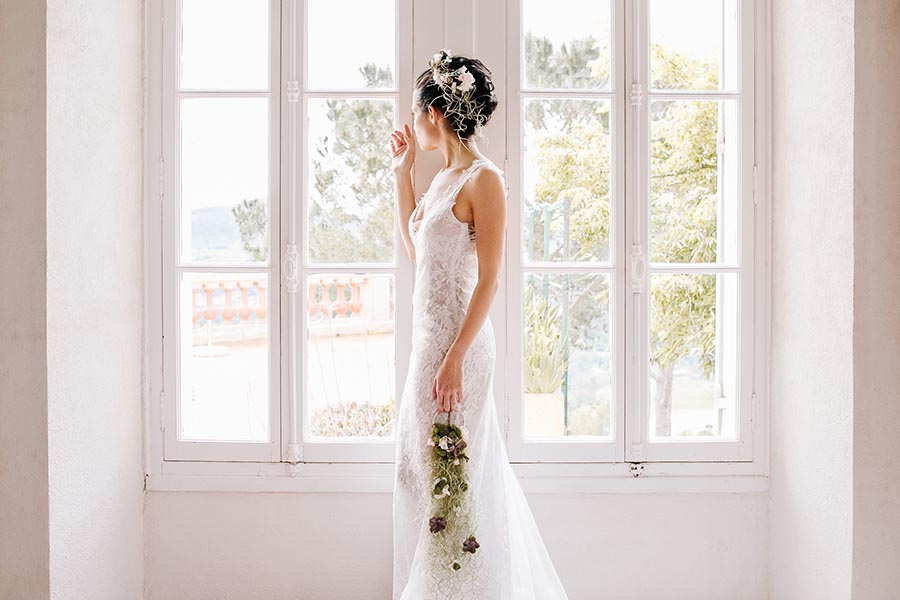 Profil d'une mariée en robe de dentelle blanche posant devant une grande fenêtre lumineuse lors de ses préparatifs à Marseille.